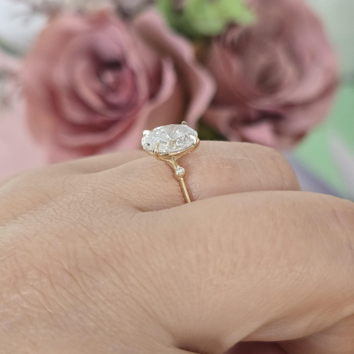 Gold ring with a large diamond on a finger, blurred pink flowers in the background