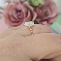 Gold ring with a large diamond on a finger, blurred pink flowers in the background